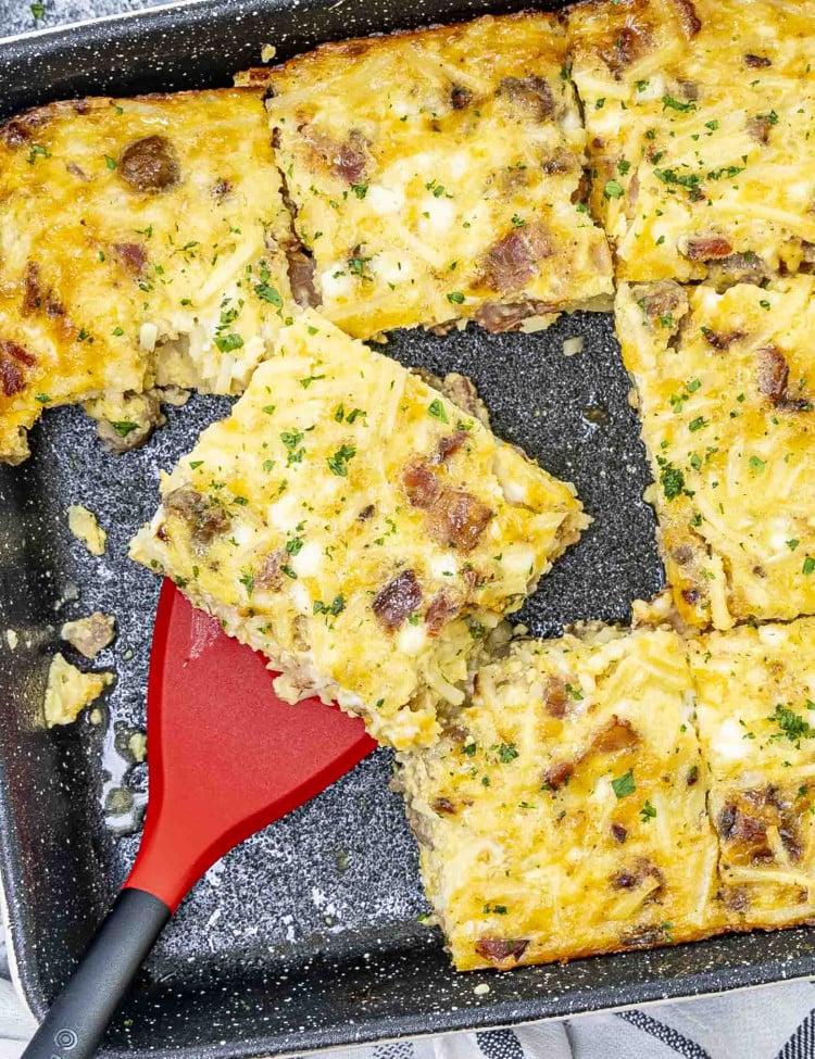 A slice of Amish Breakfast Casserole is being lifted from a baking dish with a red spatula.