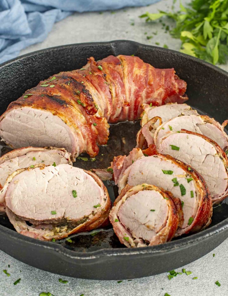 Bacon-wrapped pork tenderloin, sliced into medallions in a cast-iron skillet, garnished with fresh parsley.