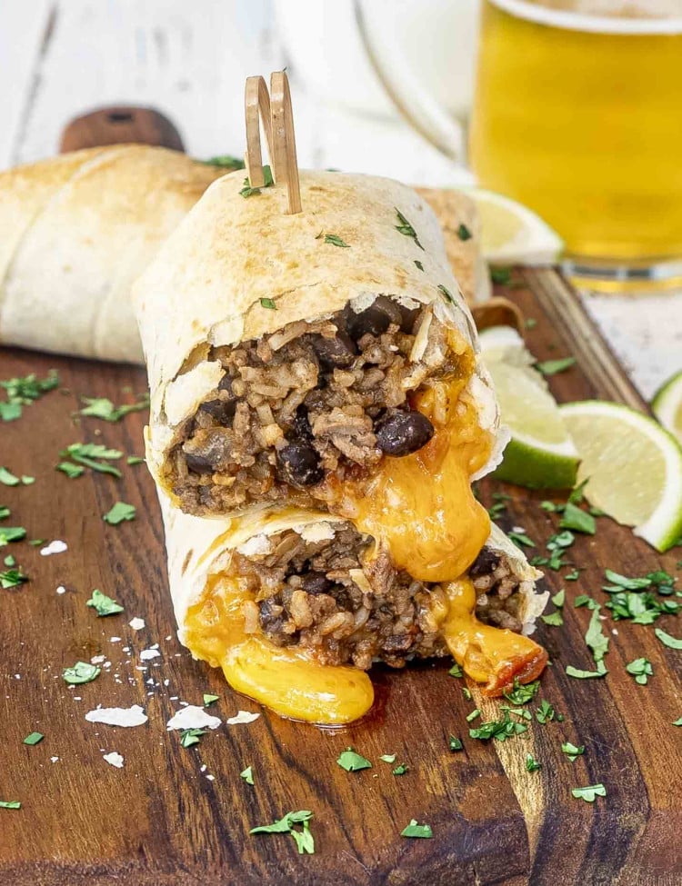 a beef burrito cut in half with melty cheese, on a cutting board with a glass of beer in the background.