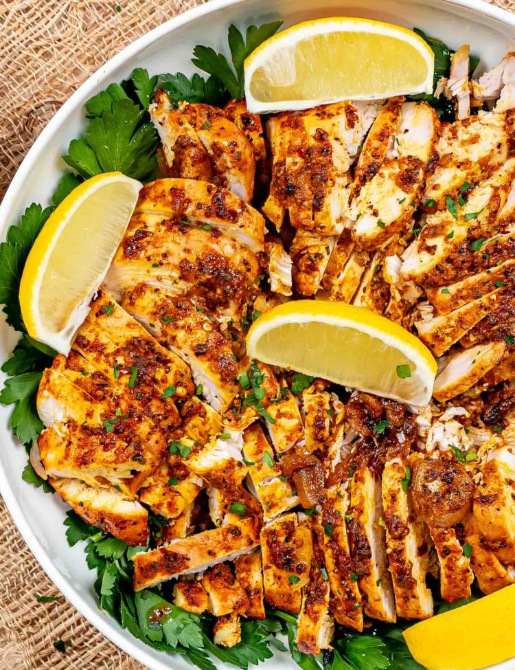 sliced chicken shawarma in a white bowl garnished with parsley and lemon wedges.