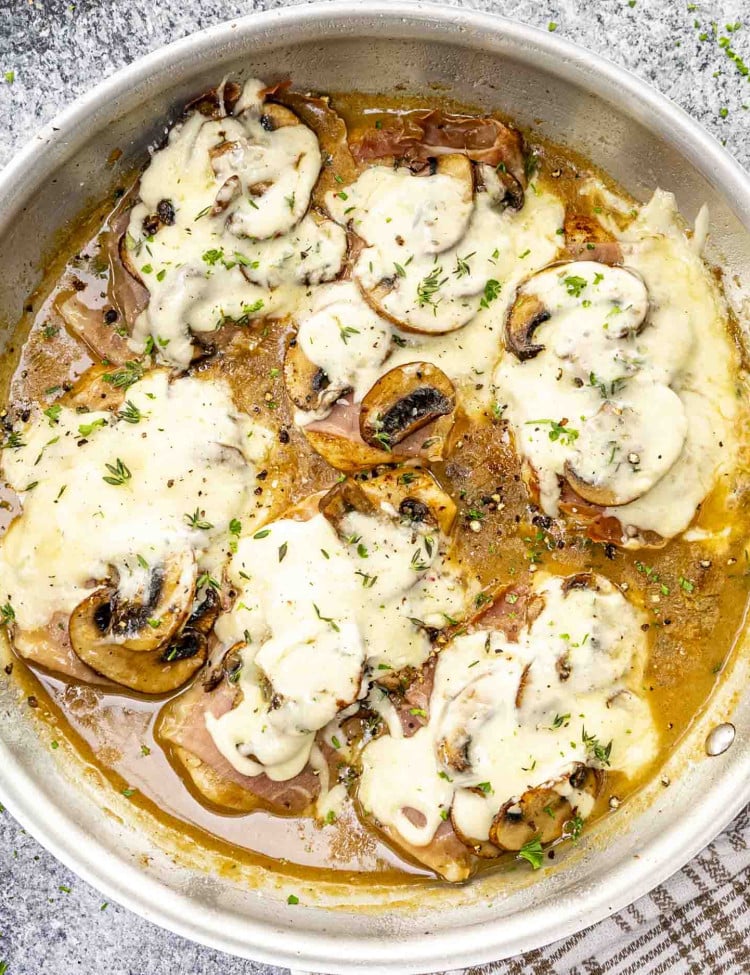 Golden chicken breasts topped with melted cheese, prosciutto, and sautéed mushrooms, served in a flavorful sauce and garnished with herbs.
