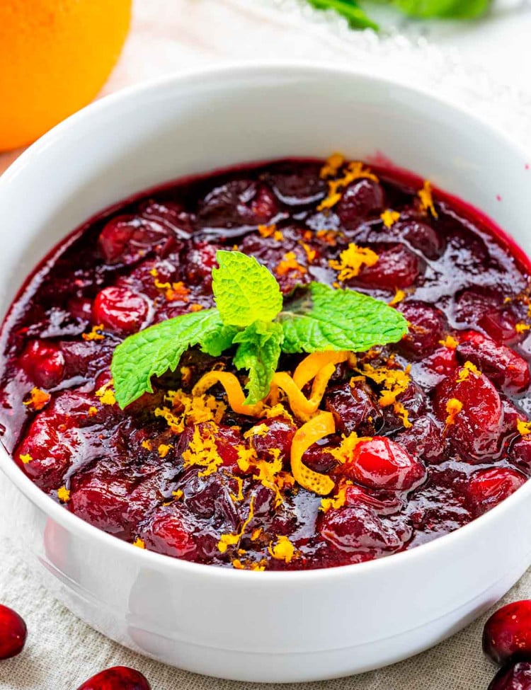 closeup of cranberry sauce in a white bowl garnished with orange zest and mint.