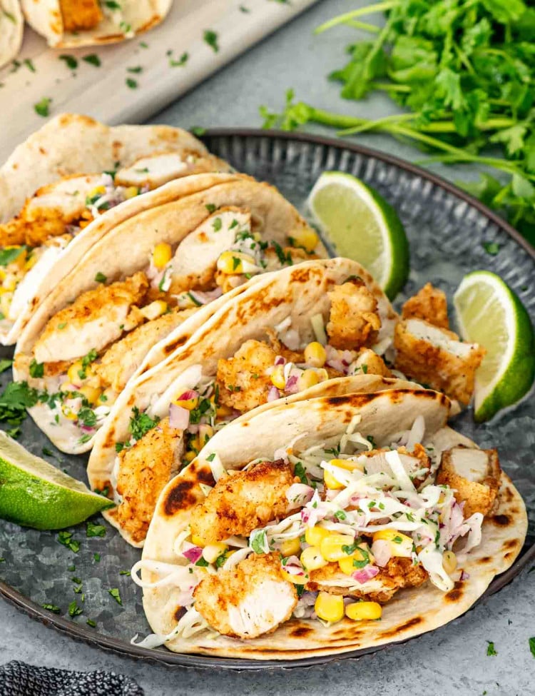 a few crispy fried chicken tacos on a metal plate garnished with lime wedges.