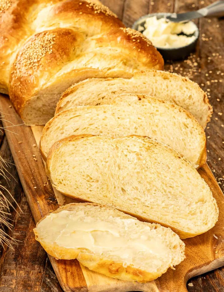 A fresh, golden-braided no knead challah bread on a wooden board with slices and butter nearby.