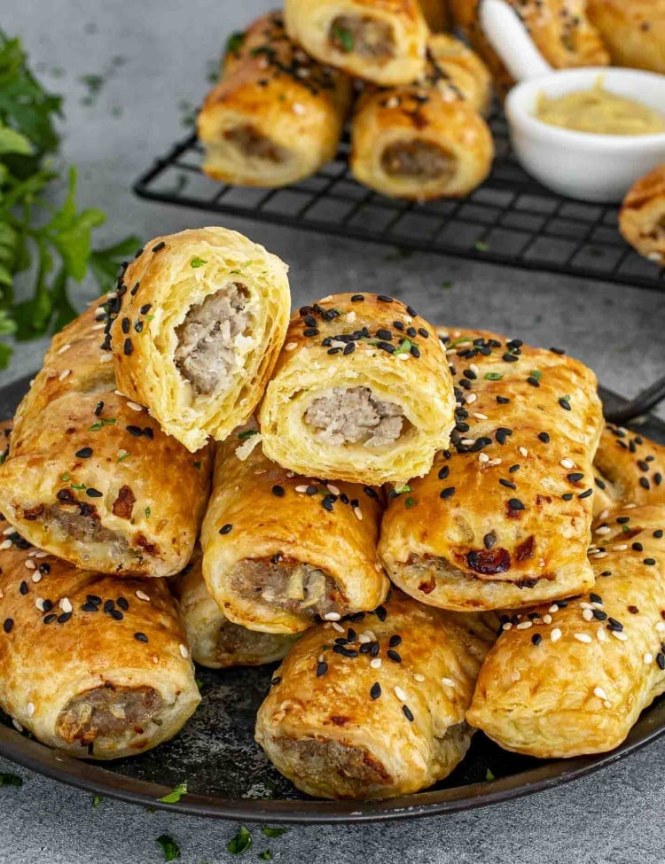 A plate stacked with golden brown pork and apple sausage rolls, topped with black and white sesame seeds. One sausage roll is cut in half, revealing the juicy sausage filling inside the flaky puff pastry. A cooling rack with more sausage rolls and a bowl of mustard are visible in the background.