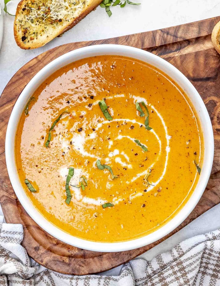 a white bowl filled with roasted red pepper soup drizzled with some cream and garnished with fresh basil.