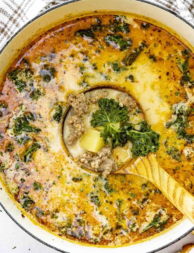 A large pot of creamy Zuppa Toscana soup filled with Italian sausage, kale, and potatoes. A ladle is scooping up a hearty portion, showing the rich broth, tender potatoes, and wilted kale.